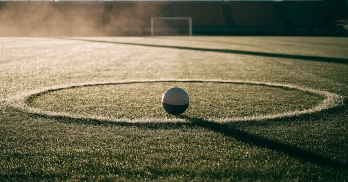 Uma bola de futebol no ponto central de um relvado, à luz da manhã